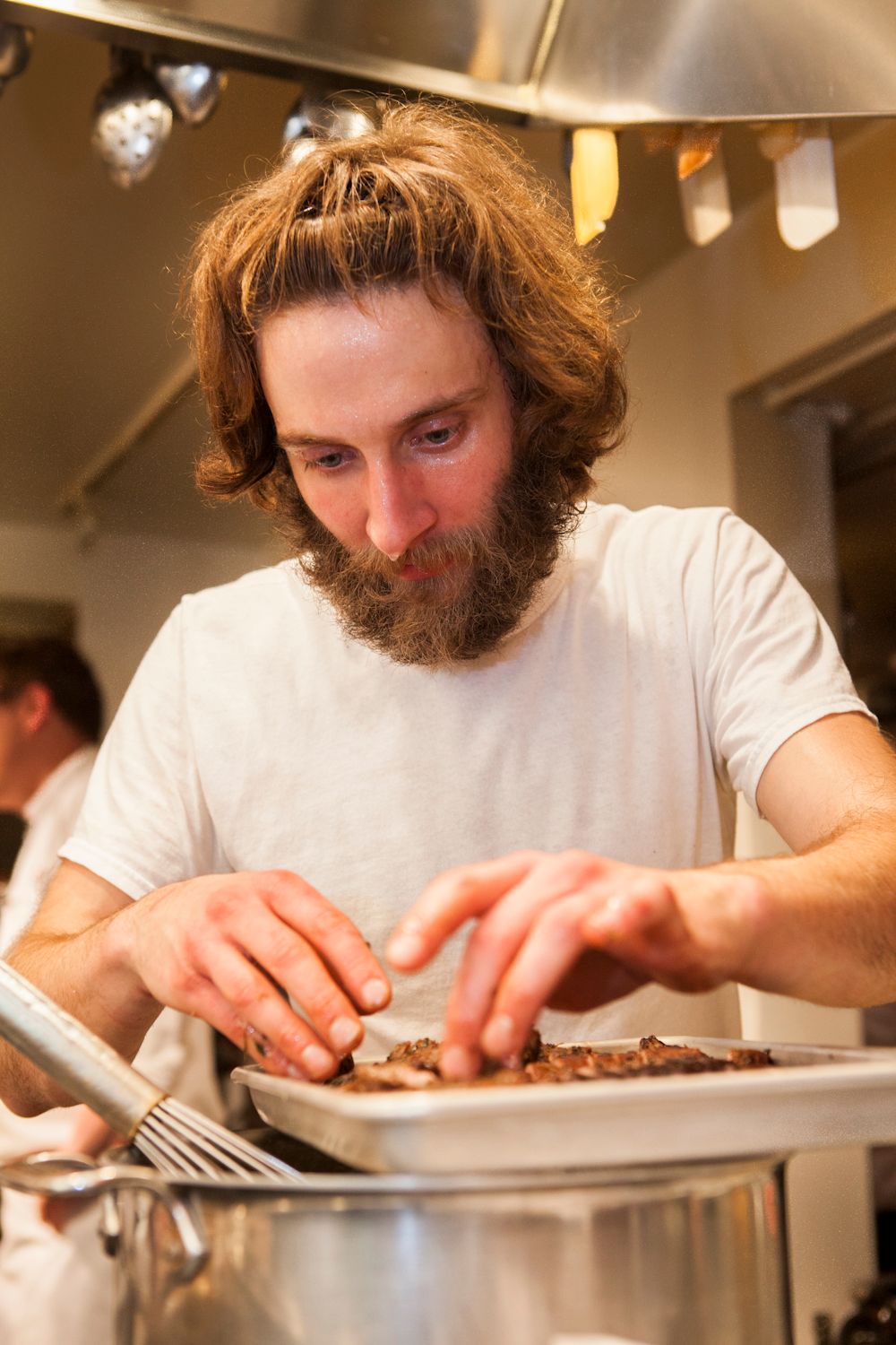 Bearded male chef carefully garnishing a tray of meat dishes in a professional kitchen.