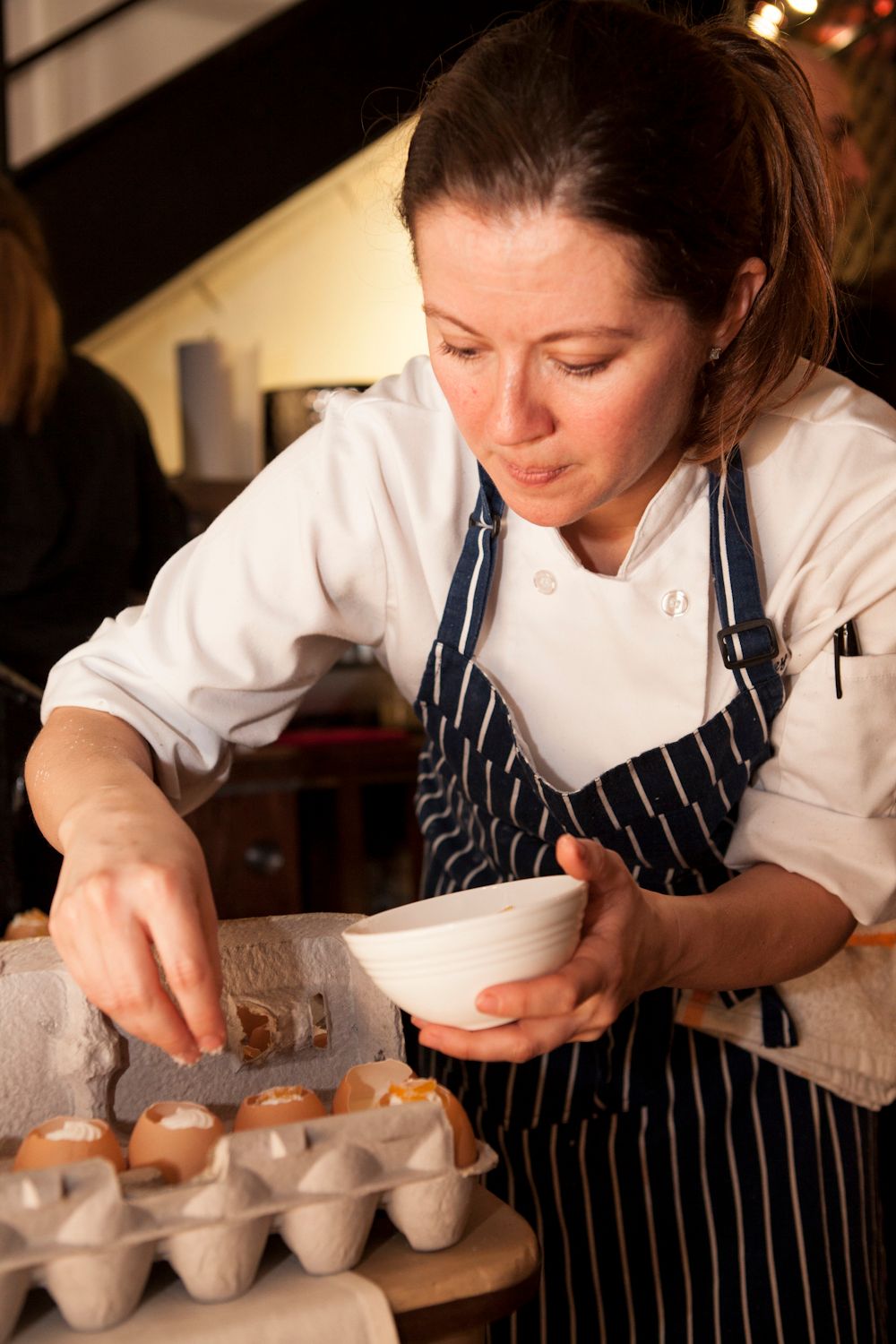 Female chef in striped apron carefully filling eggshells with gourmet mixture from a white bowl.