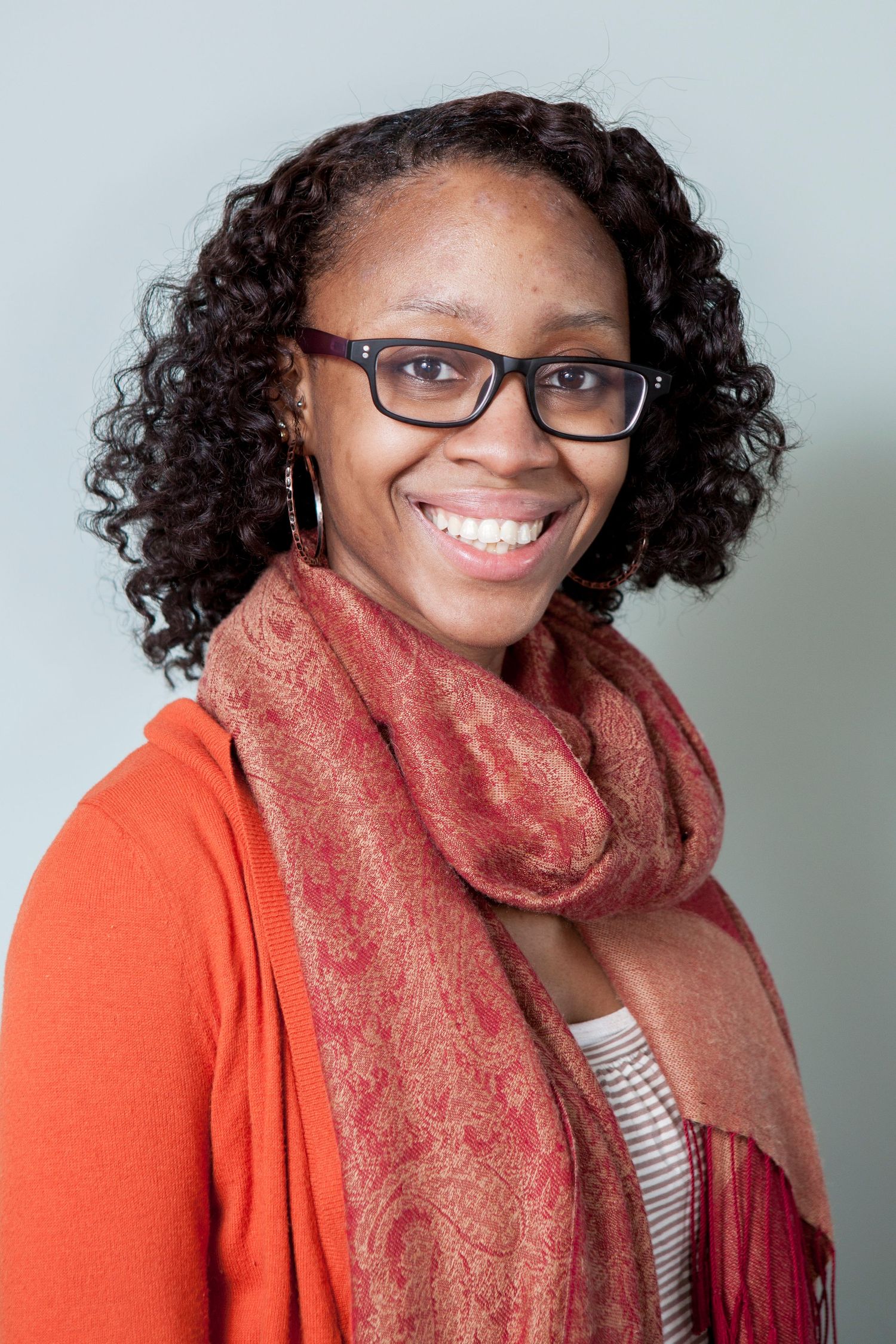 Smiling Black woman with curly hair, glasses, and a red scarf, wearing an orange cardigan.