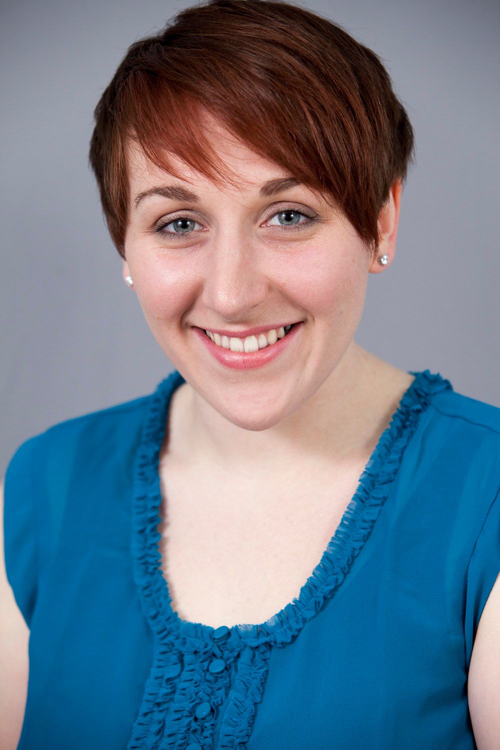 Young woman with short red hair smiling, wearing a blue ruffled top, studio headshot.