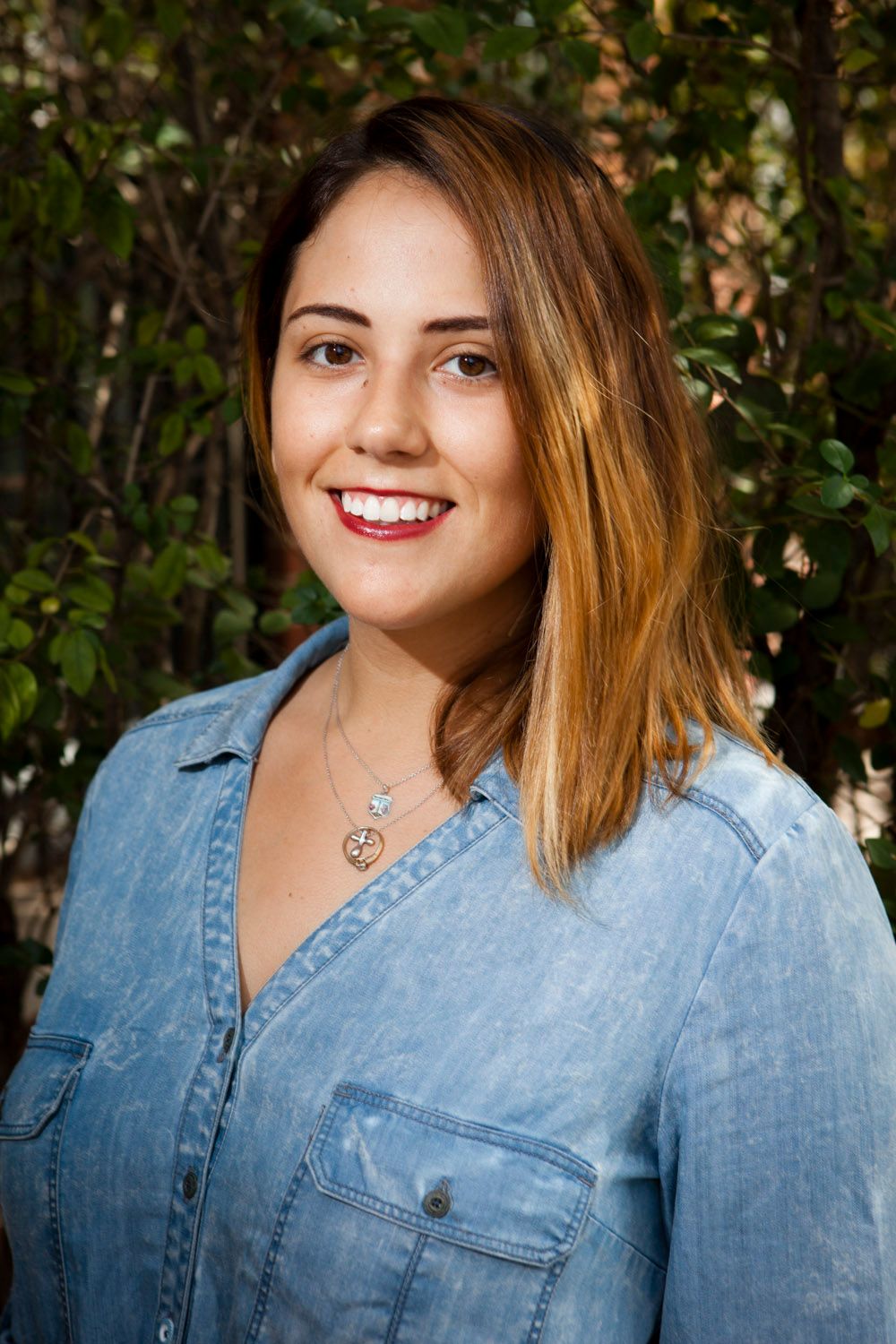 Smiling young woman in denim shirt with necklace posing outdoors near green trees.