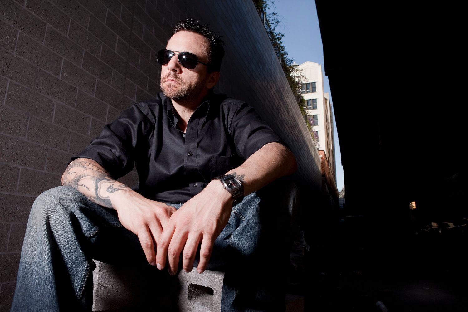 Tattooed man in black shirt and sunglasses sitting against a brick wall in an urban setting.