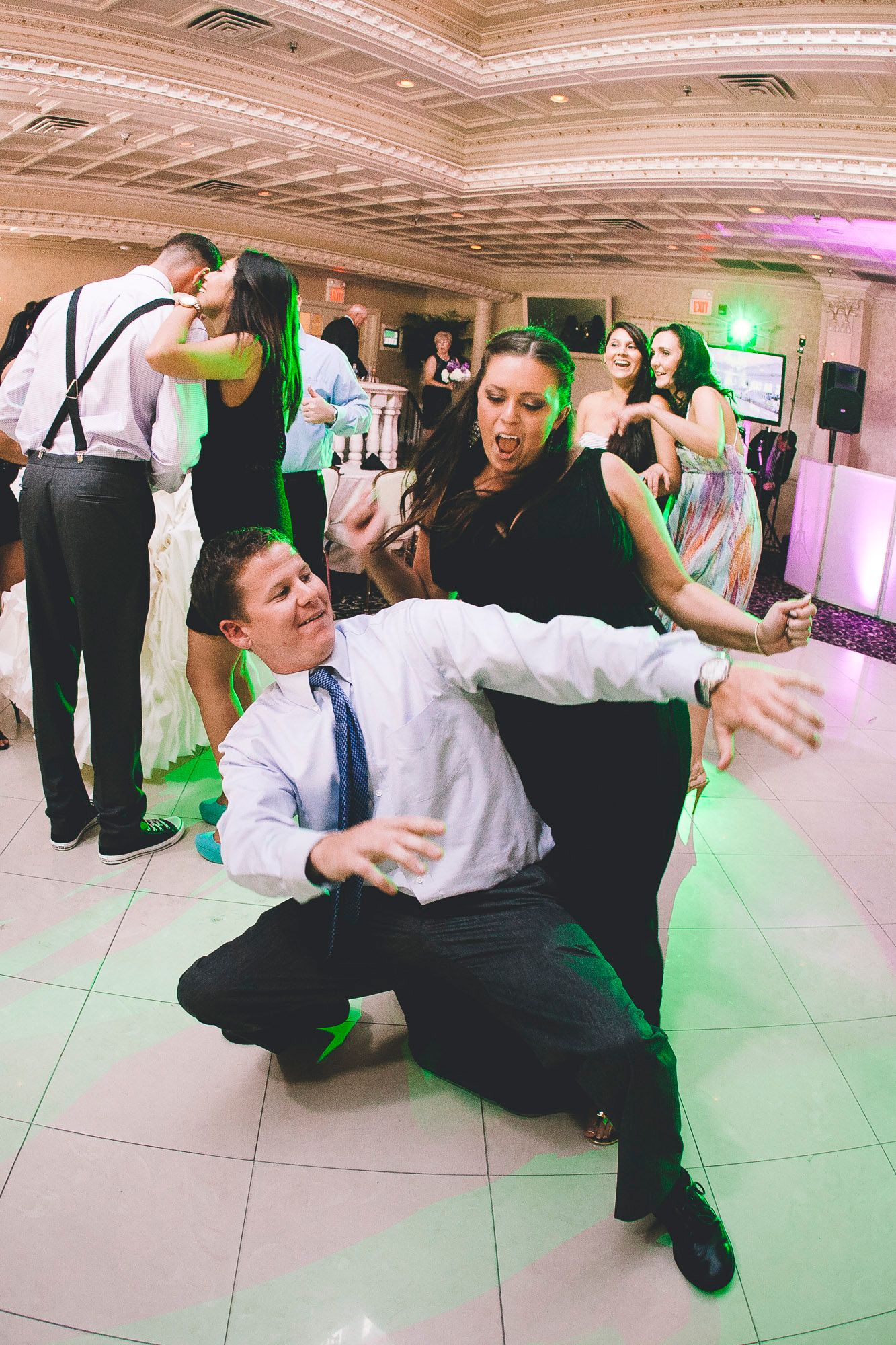 Wedding guests dancing energetically on the dance floor, with one man kneeling low under green lights.