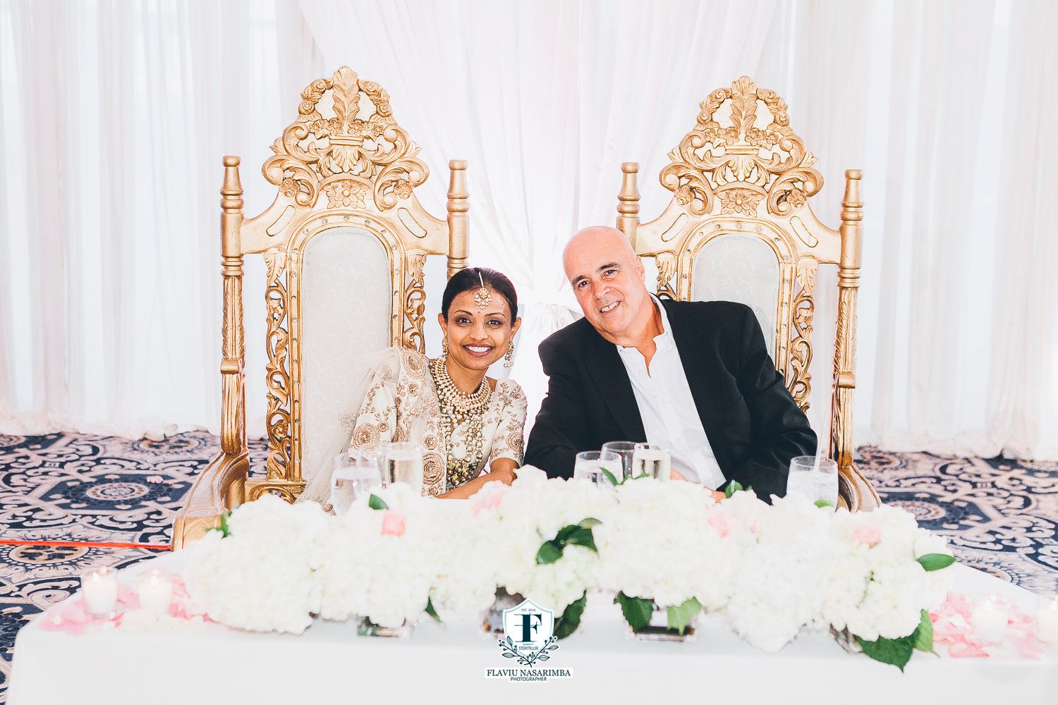 Indian bride and groom smile together seated at elegant sweetheart table adorned with white floral arrangements.