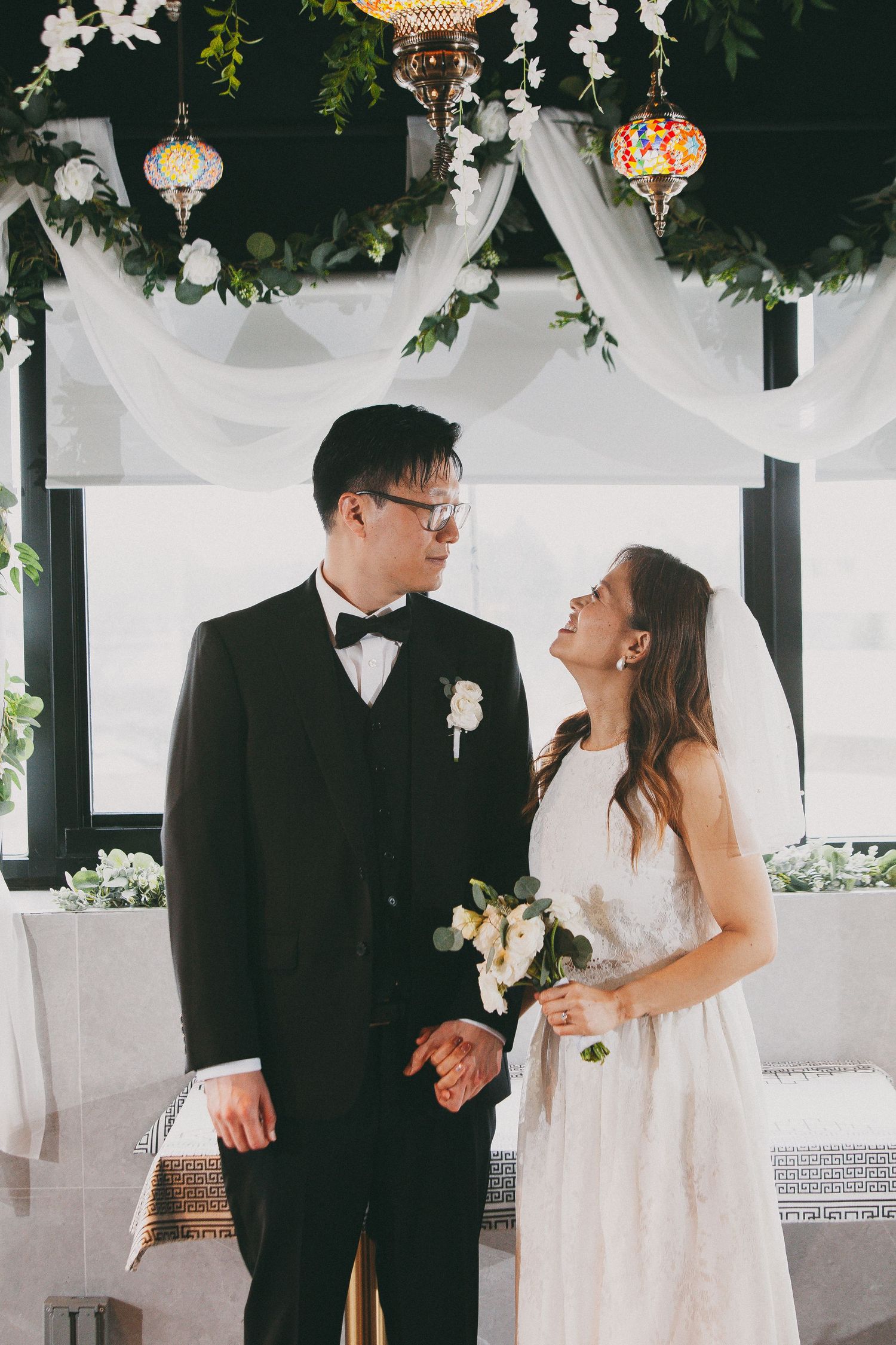 Bride and groom share a joyful moment under white draped floral arch with mosaic lanterns.