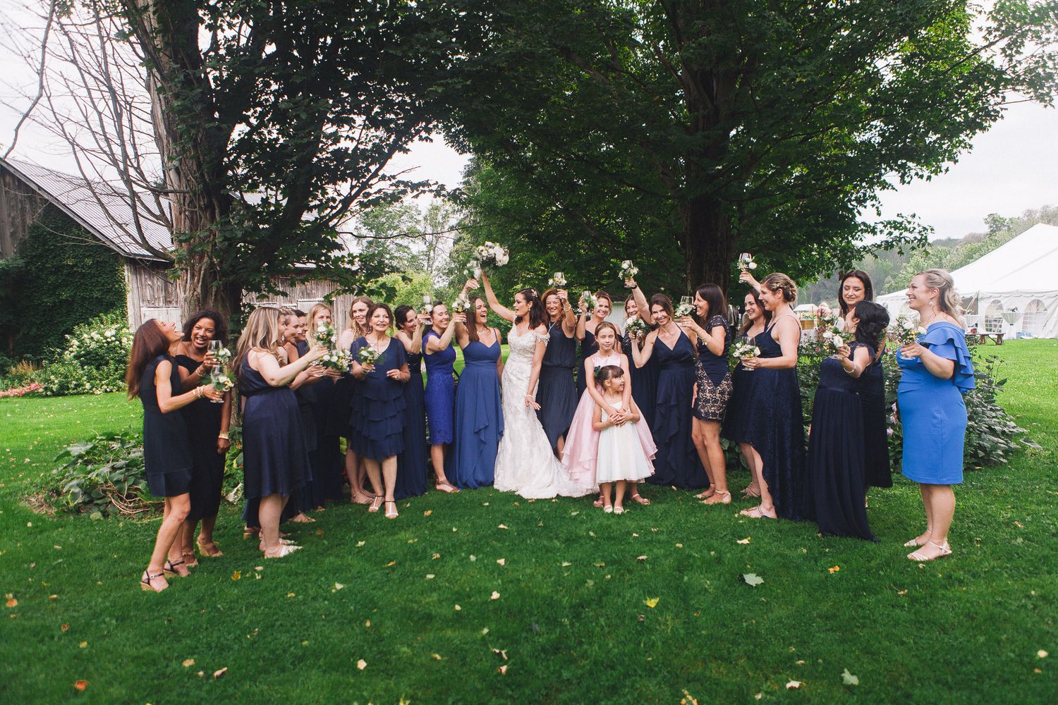 Bride surrounded by large bridal party in navy and blue dresses celebrating outdoors under trees.