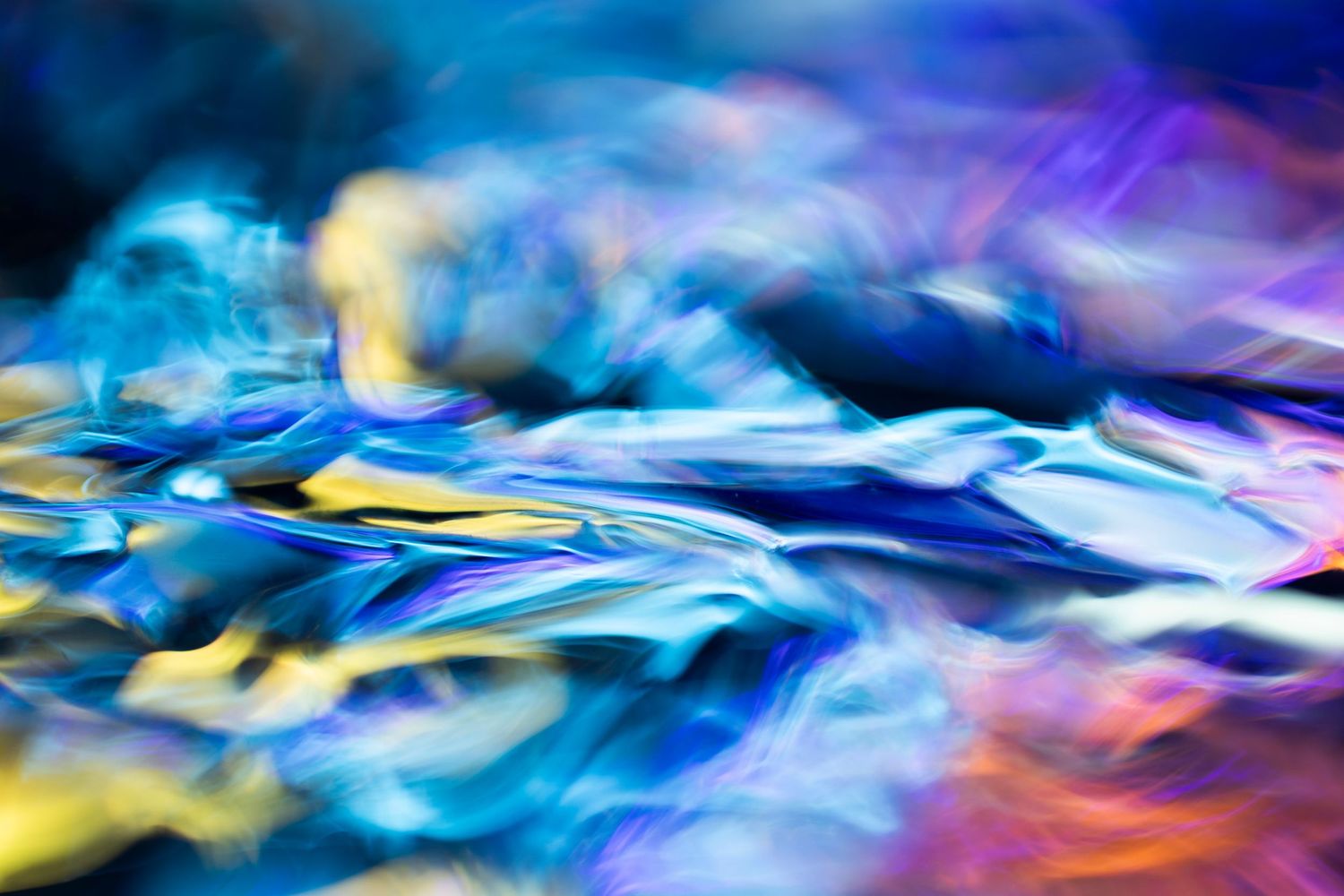 Abstract macro photograph of iridescent blue, purple, and yellow swirling light patterns creating a vibrant, fluid texture.