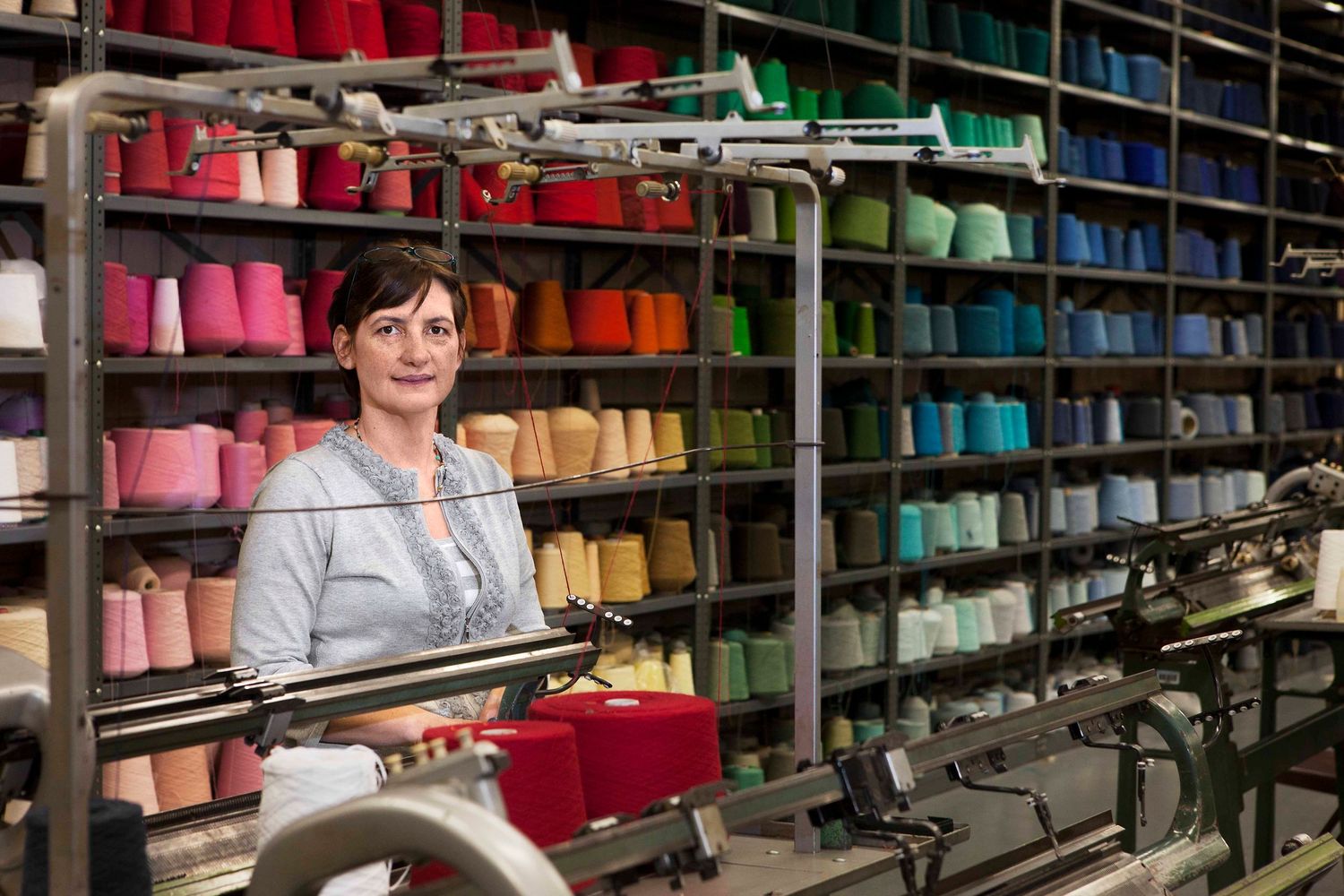 Smiling woman standing in a textile factory surrounded by colorful yarn spools on shelves.