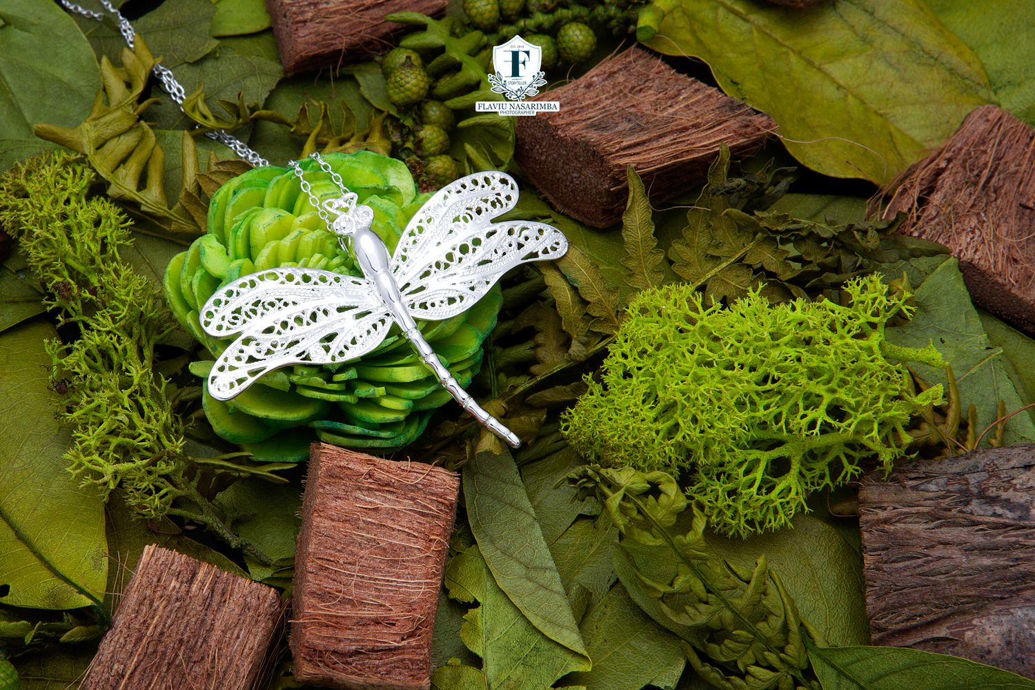 Elegant silver dragonfly pendant necklace displayed on a bed of green leaves and bark.