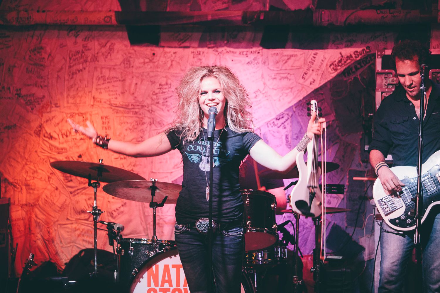 Blonde female singer performs energetically on stage with band and drum kit visible behind her.