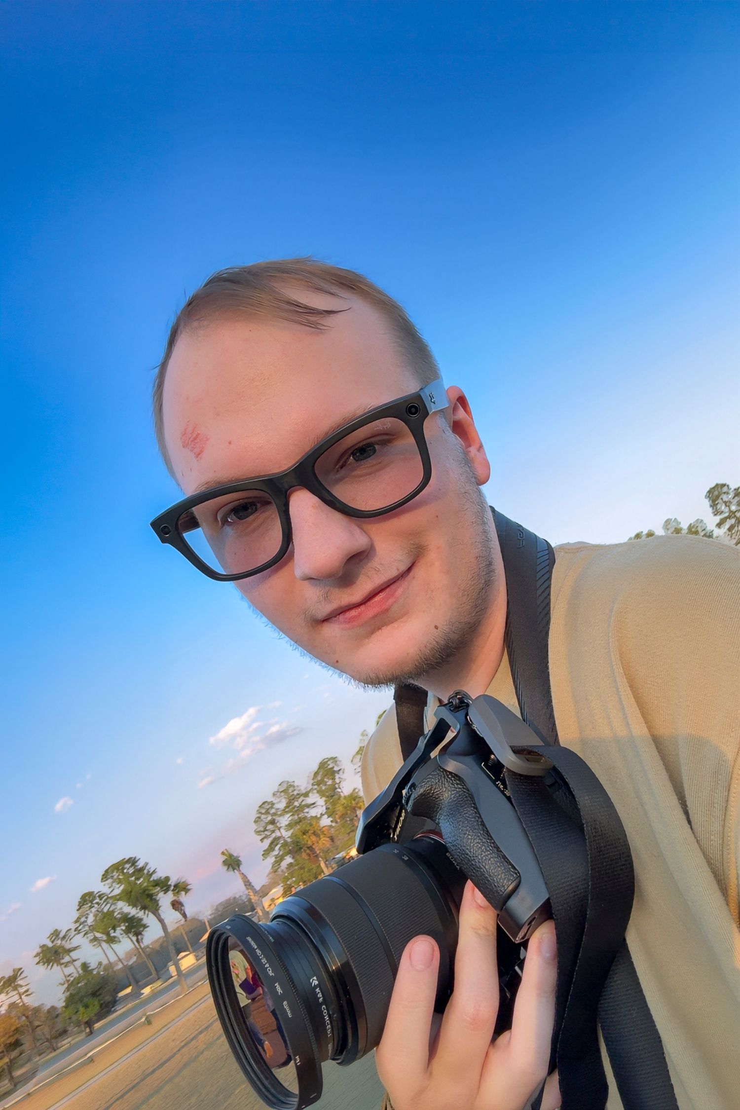 A young man with glasses holds a DSLR camera outdoors under a clear blue sky.
