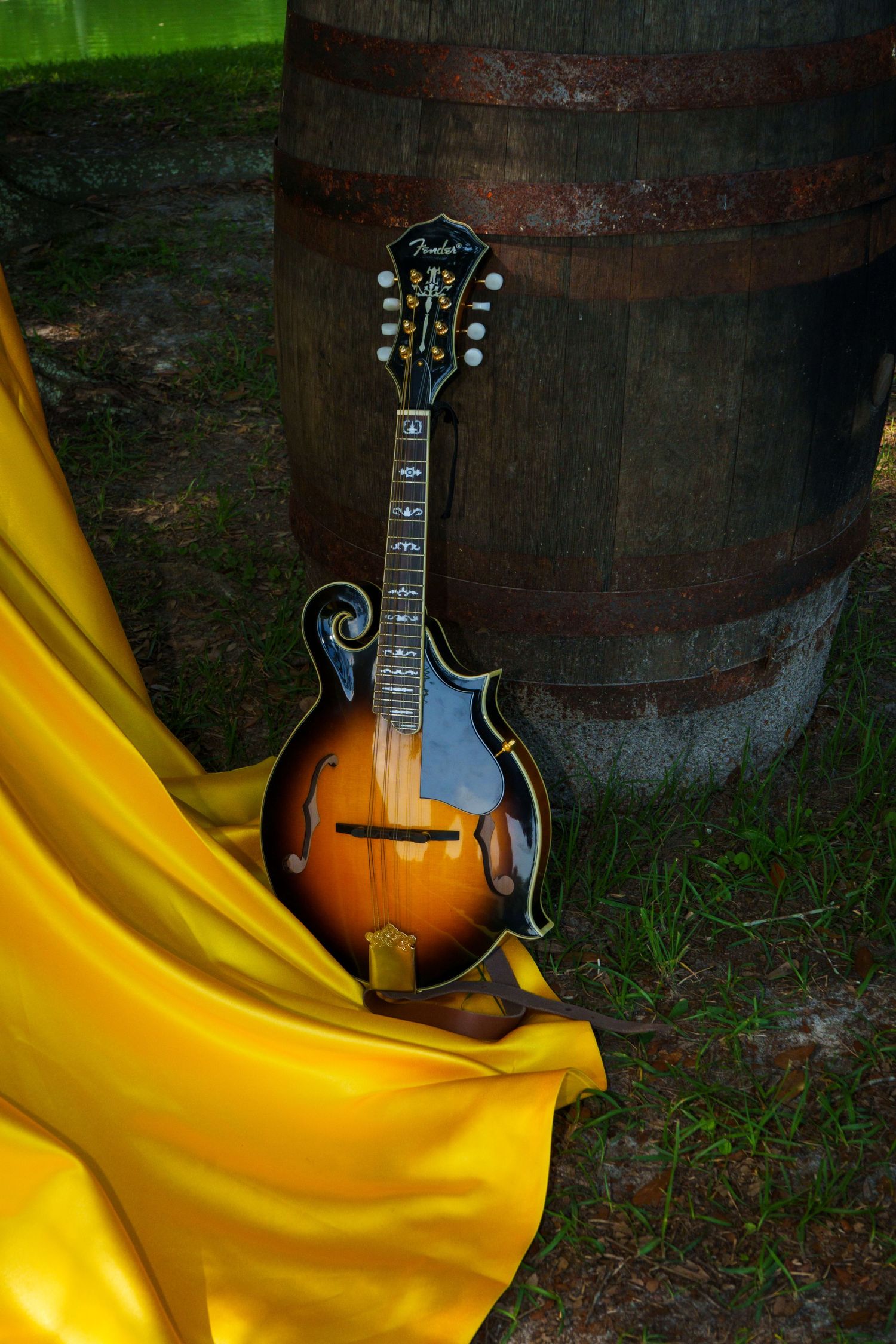 A Fender mandolin leaning against a wooden barrel with yellow satin fabric outdoors.