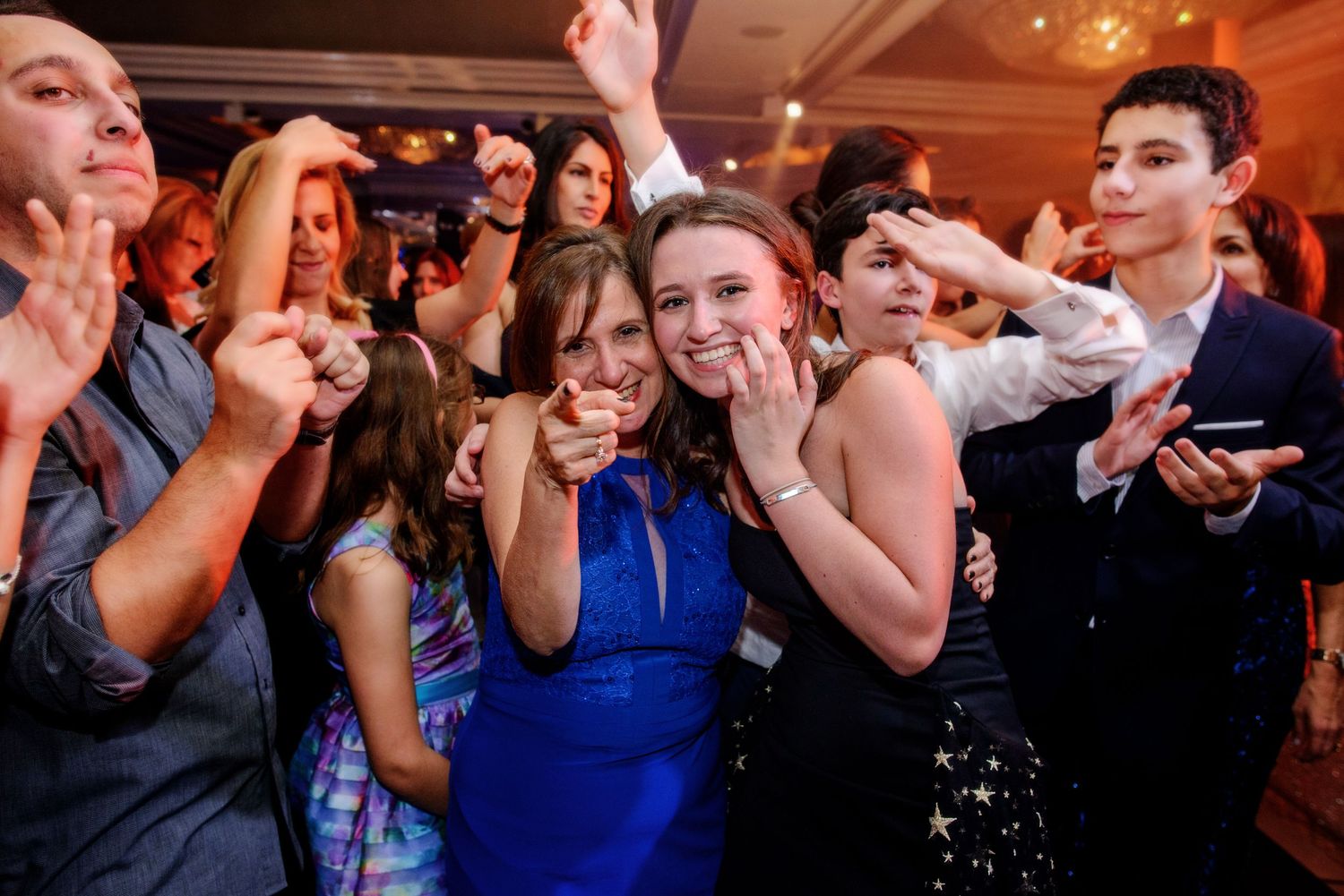 Wedding guests dancing and celebrating on a crowded dance floor at an indoor reception venue with warm lighting.