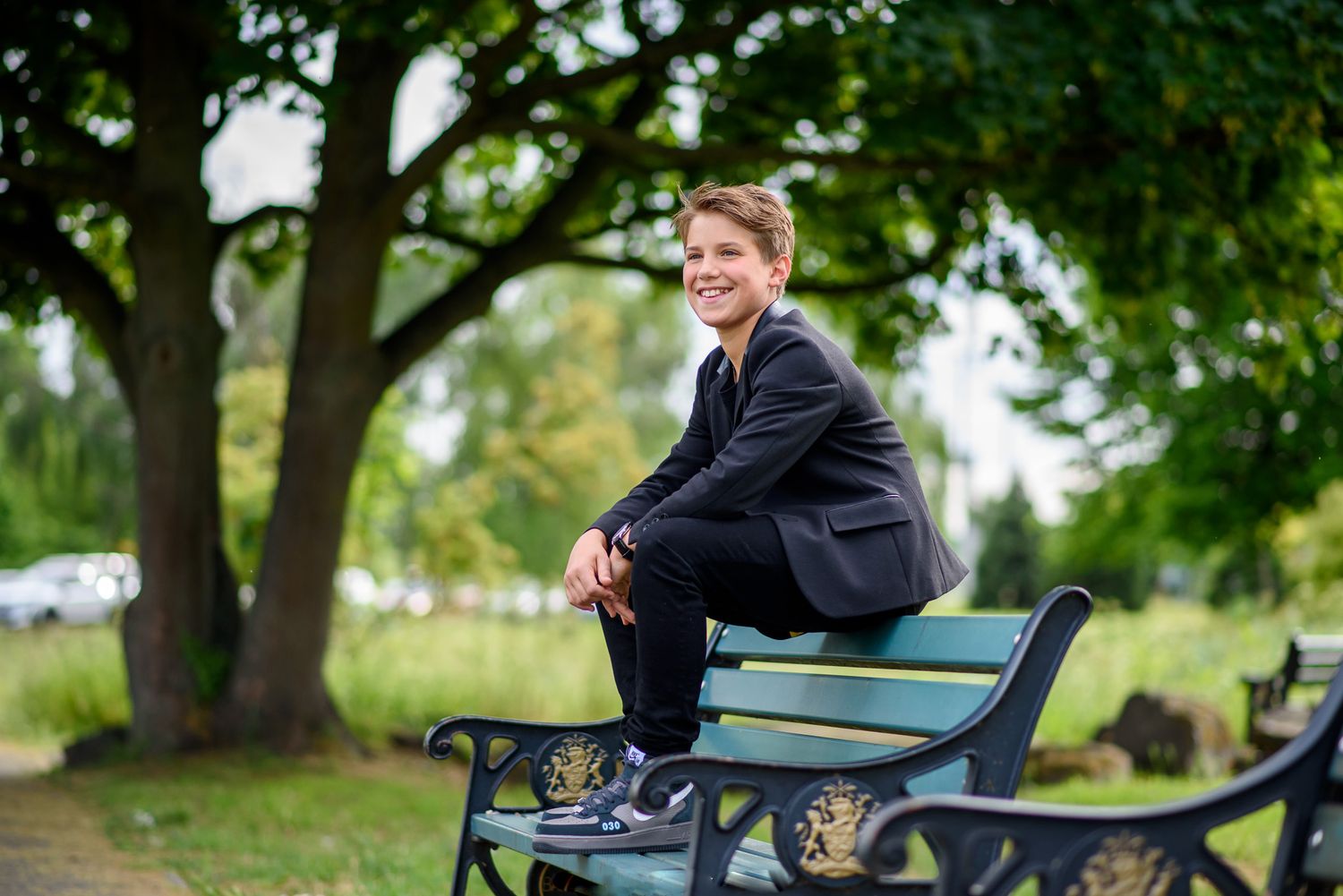A person in a blue shirt sits on a park bench surrounded by lush green trees on a sunny day.