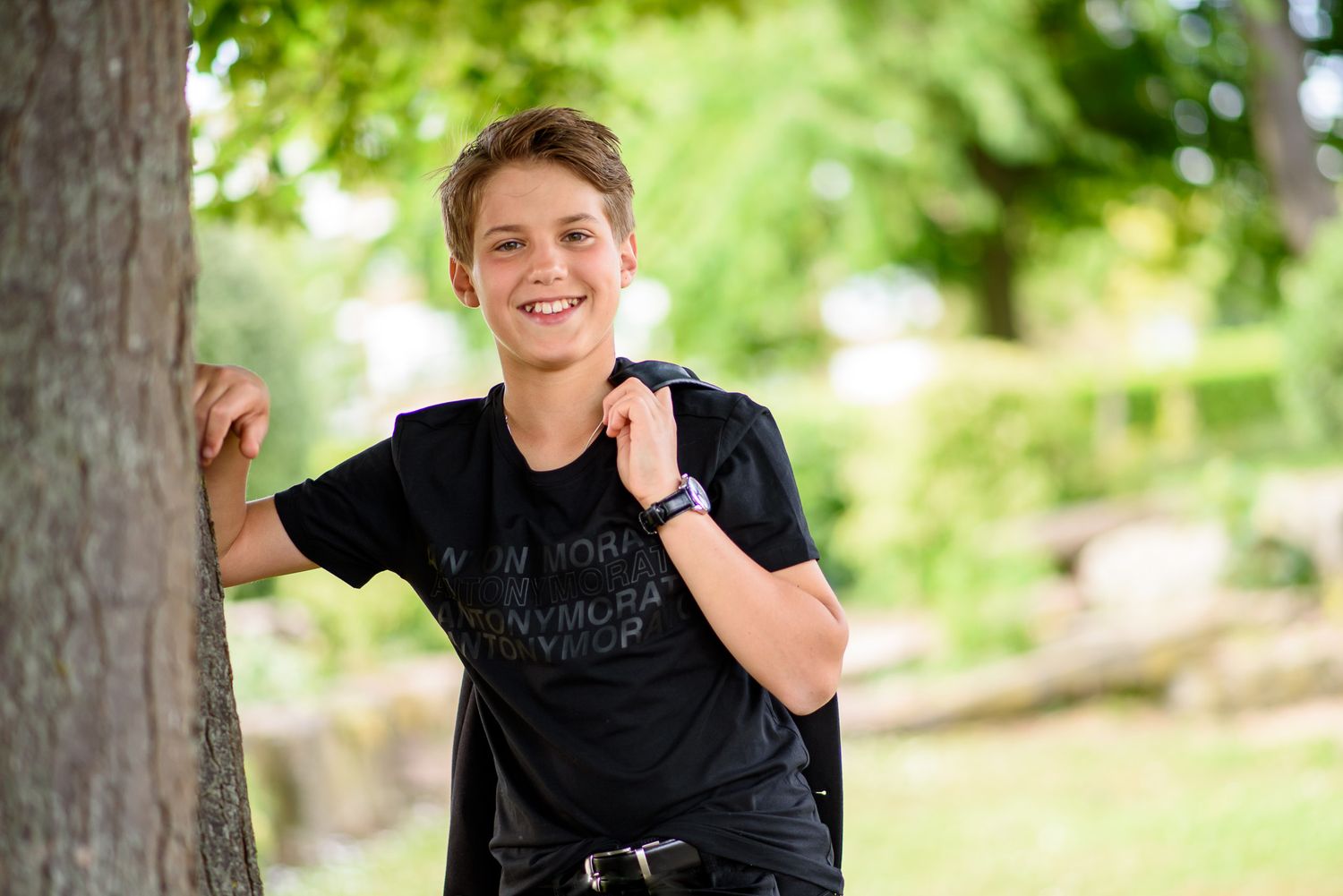 Casual portrait of someone wearing a black t-shirt leaning against a tree in a sunny outdoor setting.