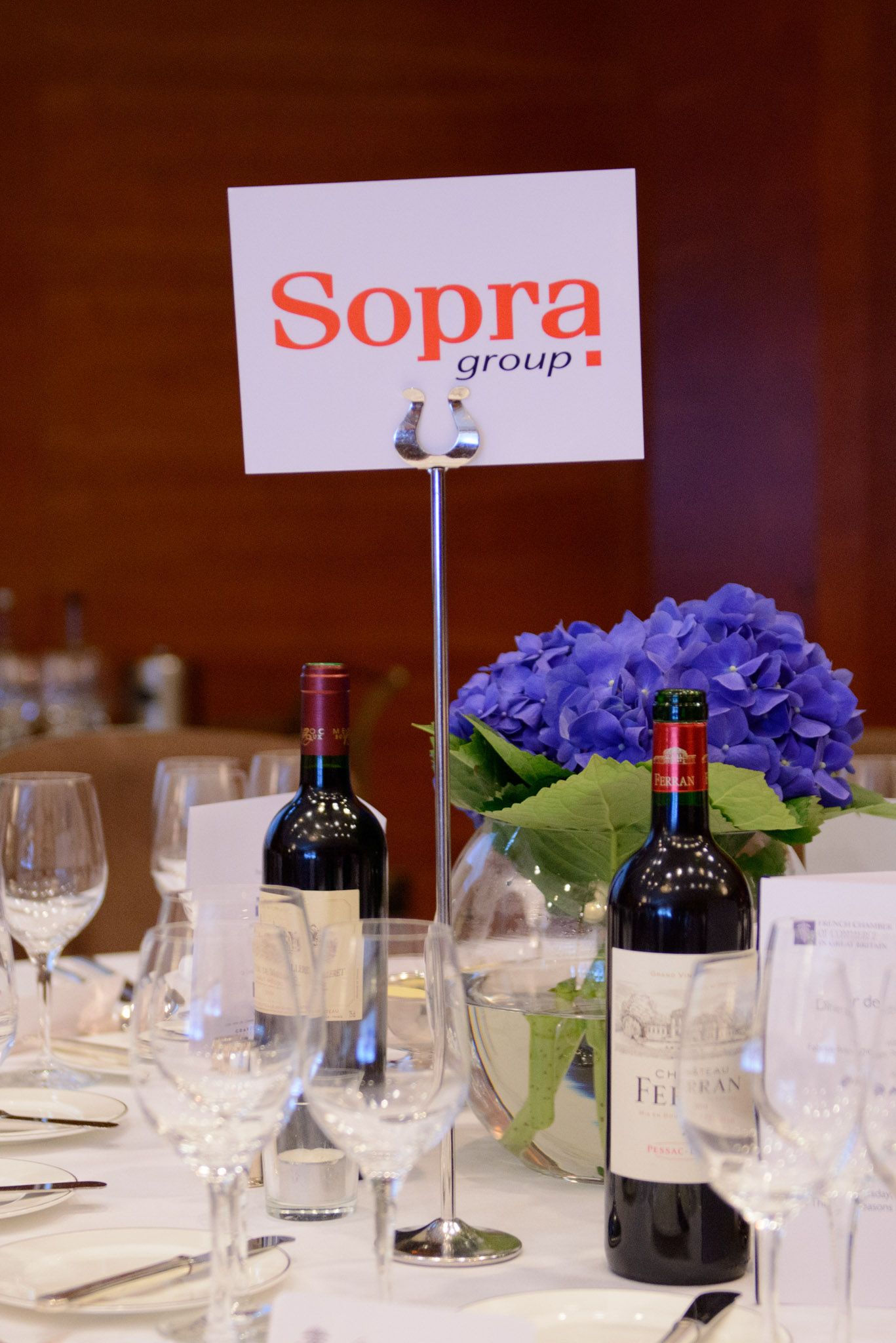 Corporate dinner table setting with Sopra Group signage, wine bottles and purple hydrangea centerpiece.