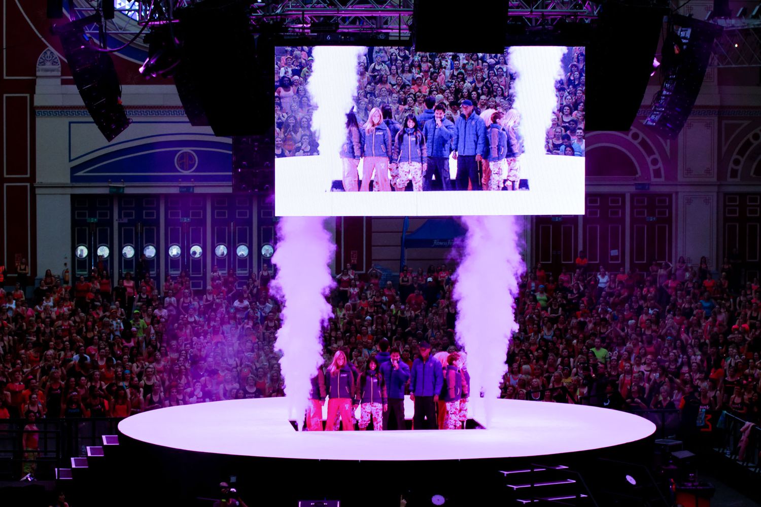Concert arena with purple stage lighting and large video screens displaying performance.