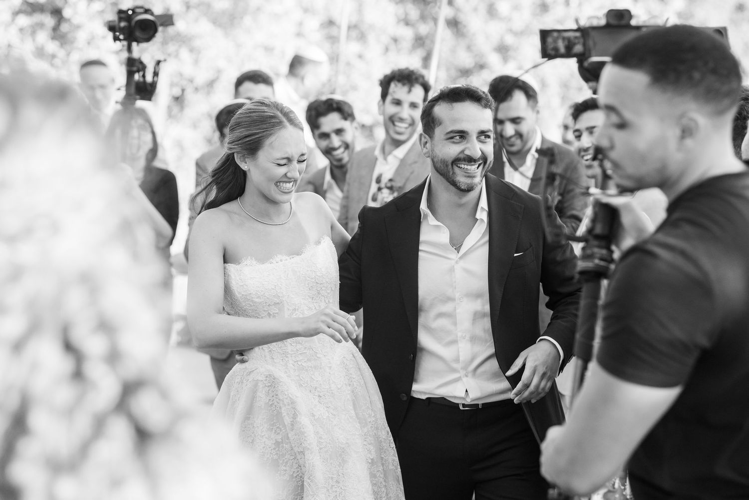 A black and white candid wedding moment showing joyful laughter and celebration among a group at an outdoor gathering.