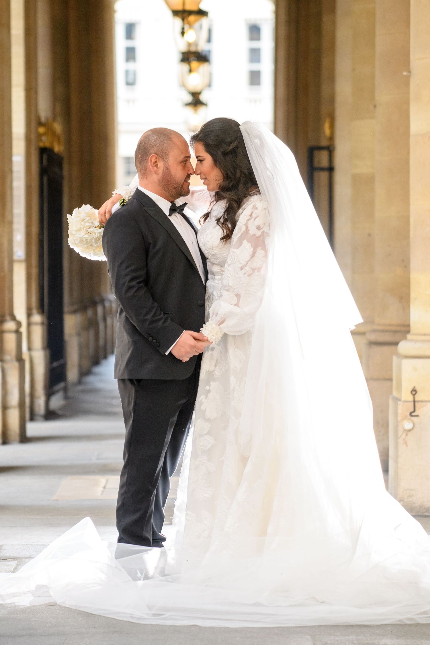 Romantic wedding couple embraces in an elegant colonnade hallway with flowing white veil and formal attire.