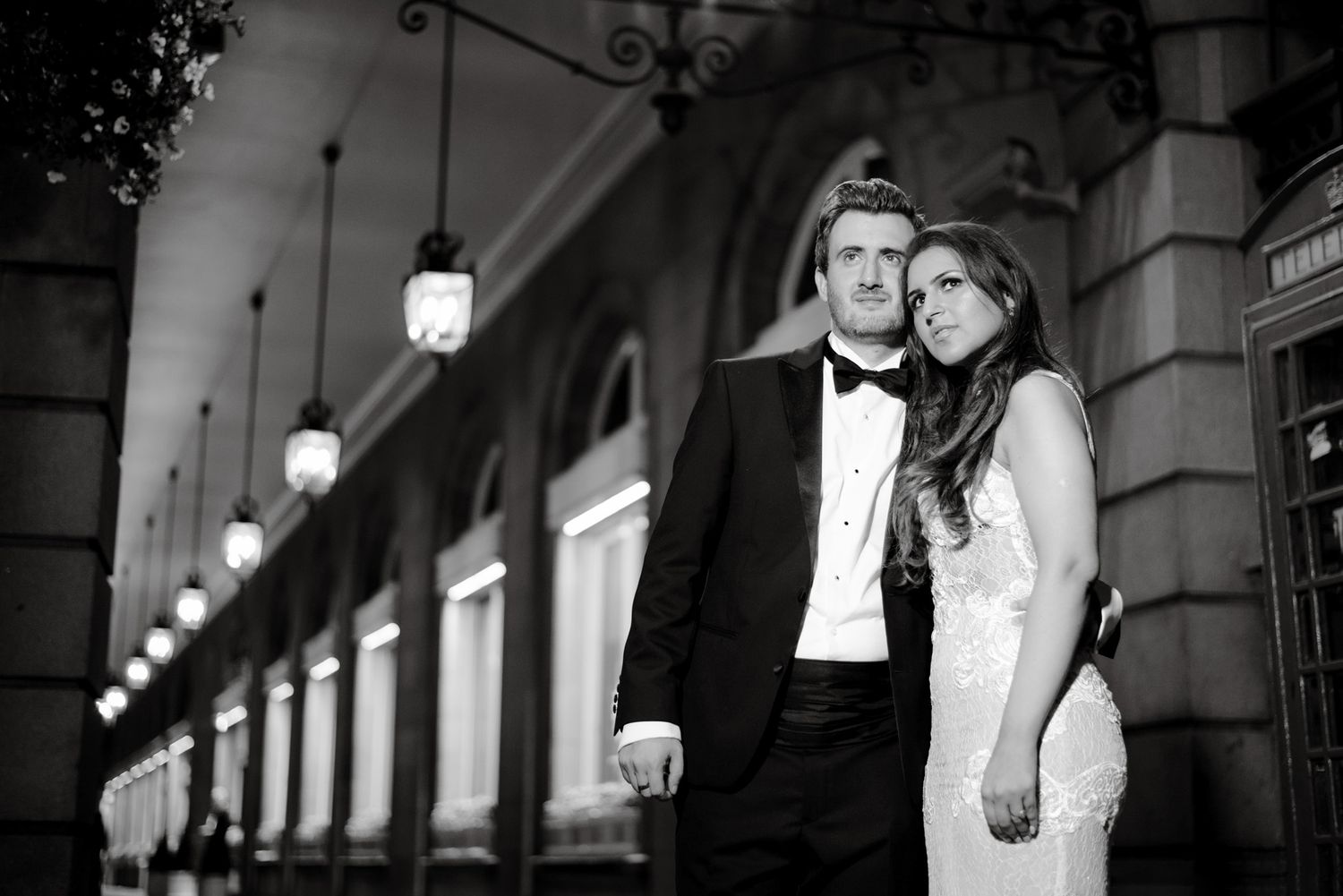 Elegant couple in formal attire posing outside a classic building with lanterns in a black and white wedding photo.