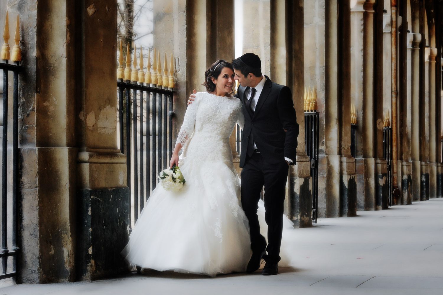 Bride and groom share a romantic moment in a grand columned hallway, bride in white gown holding bouquet.