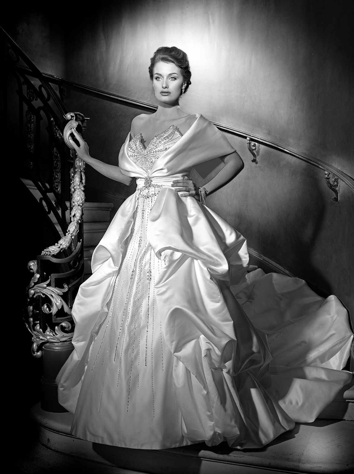 Elegant woman in a stunning white satin ball gown poses gracefully on a grand staircase in a classic black and white photo.
