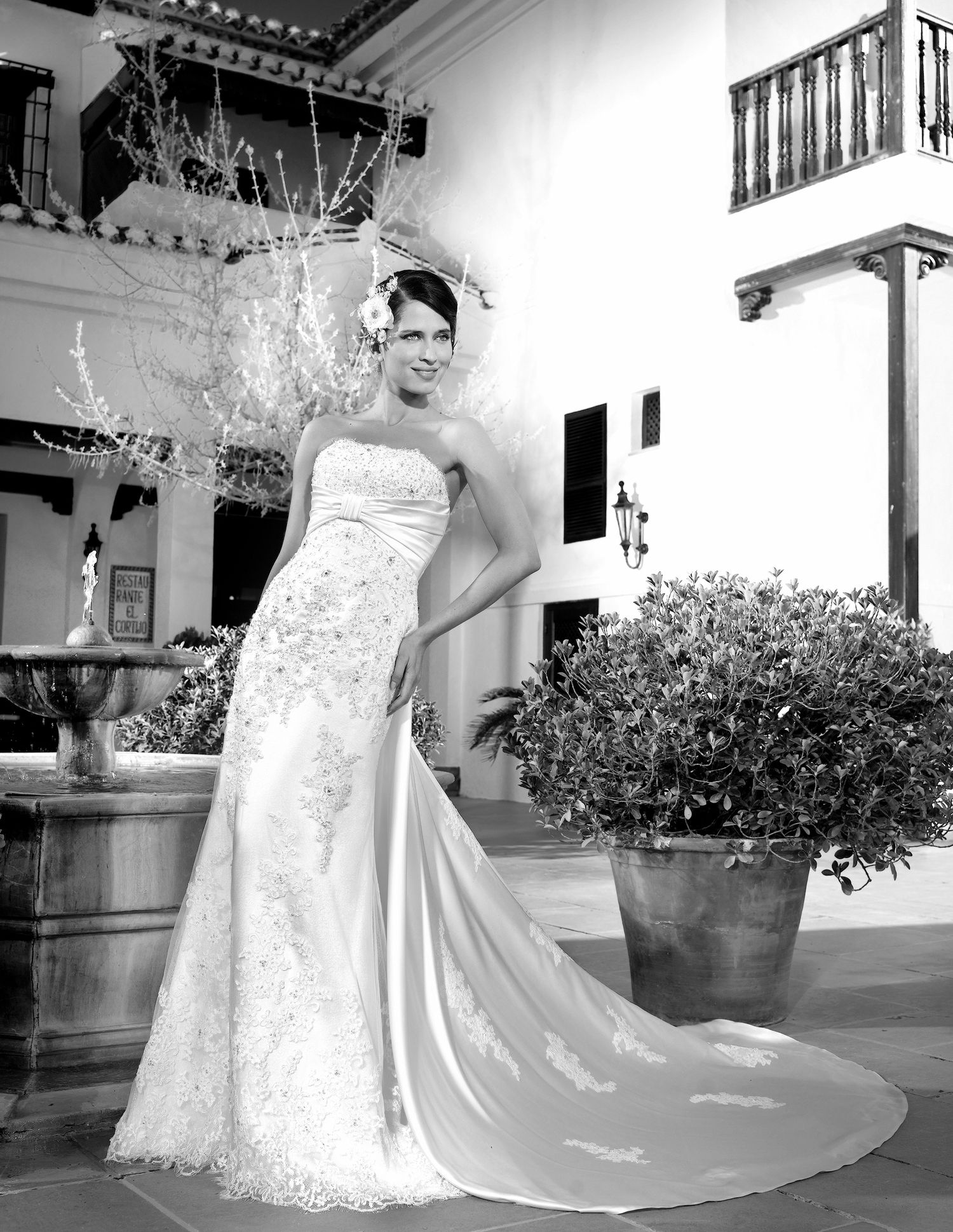 Bride in strapless lace wedding gown with train poses outdoors near fountain and large potted shrub in black and white photo.
