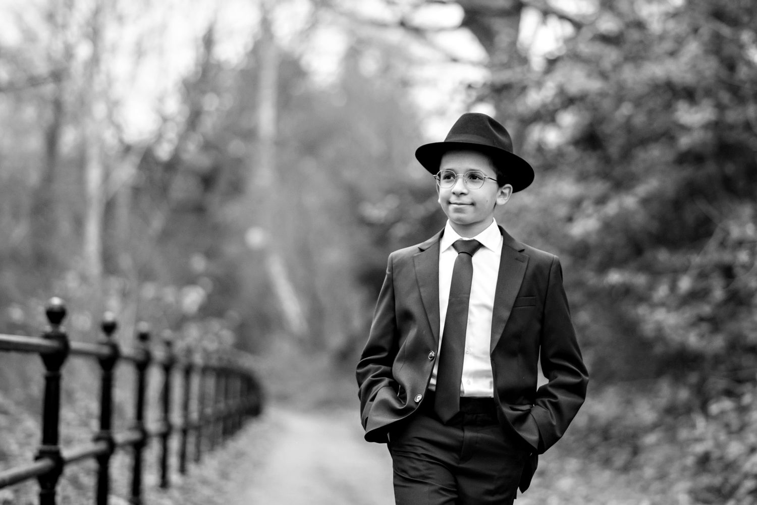 Black and white portrait of someone in a suit and fedora hat walking along an iron railing path in a park setting.