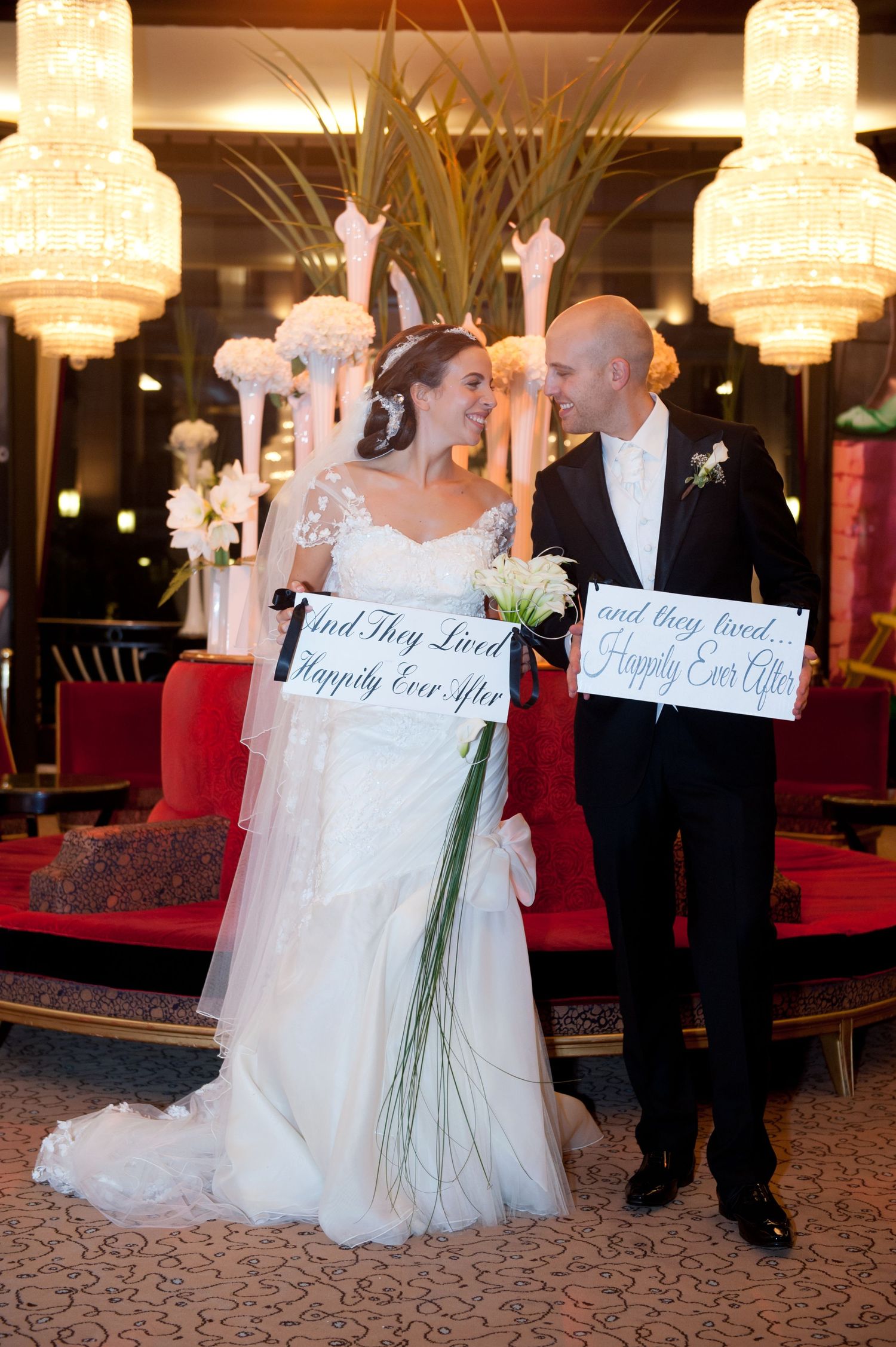 A bride and groom hold signs at their wedding reception under elegant crystal chandeliers.