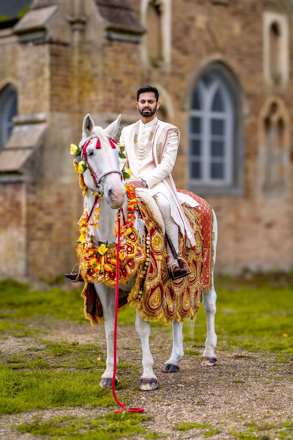 Indian groom in white sherwani riding a decorated white horse at a traditional Baraat wedding ceremony.