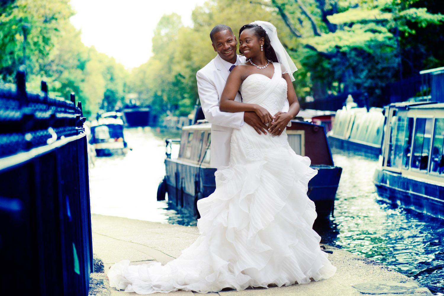Happy couple in wedding attire embracing by a scenic canal with boats in the background.