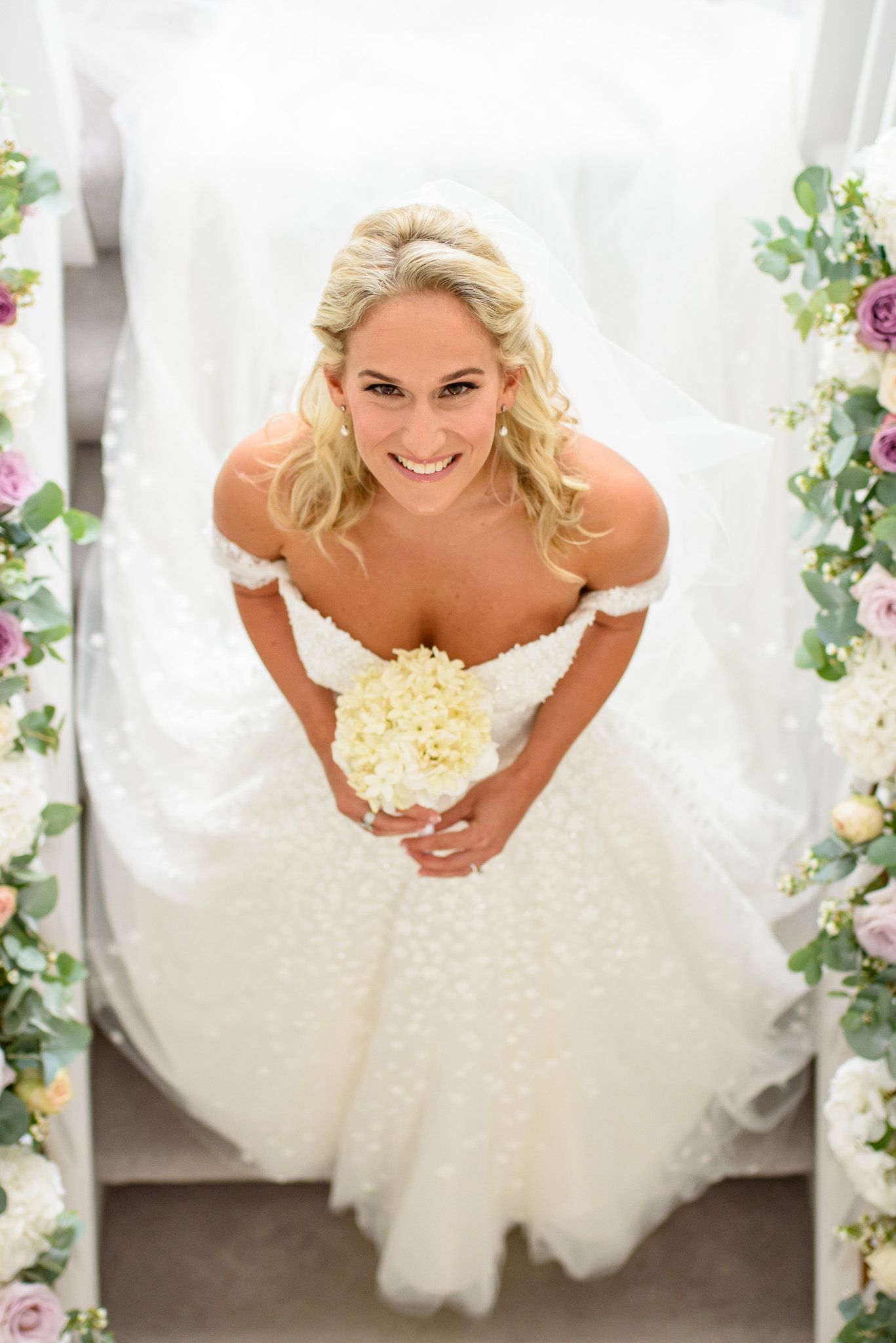 Smiling blonde bride in off-shoulder white ballgown holds cream bouquet, photographed from above between floral arrangements.