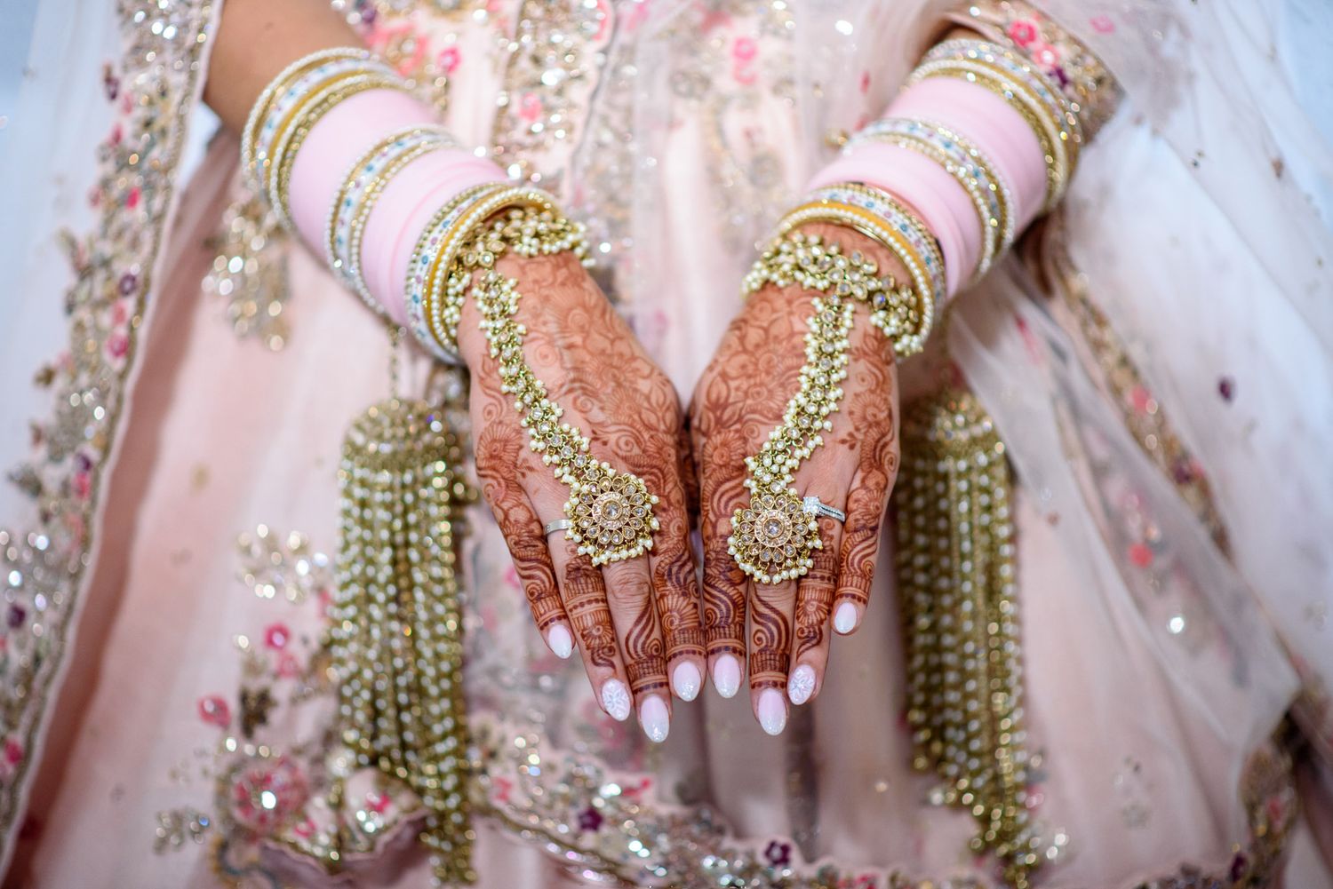 Bride's hands adorned with intricate henna, gold jewelry, bangles, and rings on a pink embroidered lehenga.