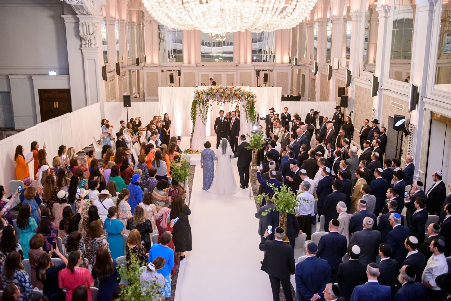 Large crowd of people gathered in an elegant white venue with crystal chandeliers for a special event.