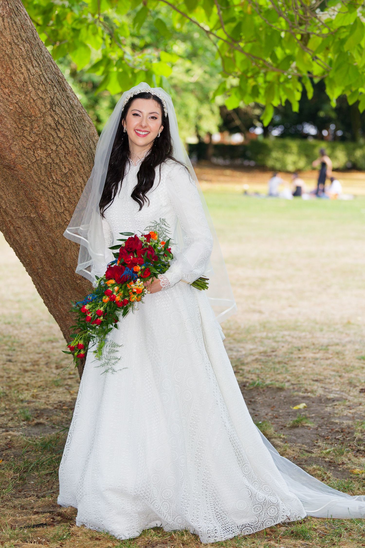 A bride in a white lace wedding dress holds a vibrant red and orange bouquet while posing outdoors under trees.