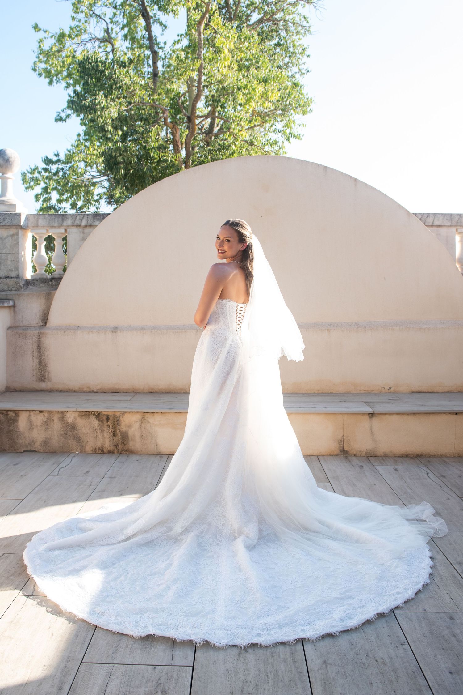 A flowing white wedding dress with intricate lace details and a dramatic cathedral train spreads across a stone terrace.