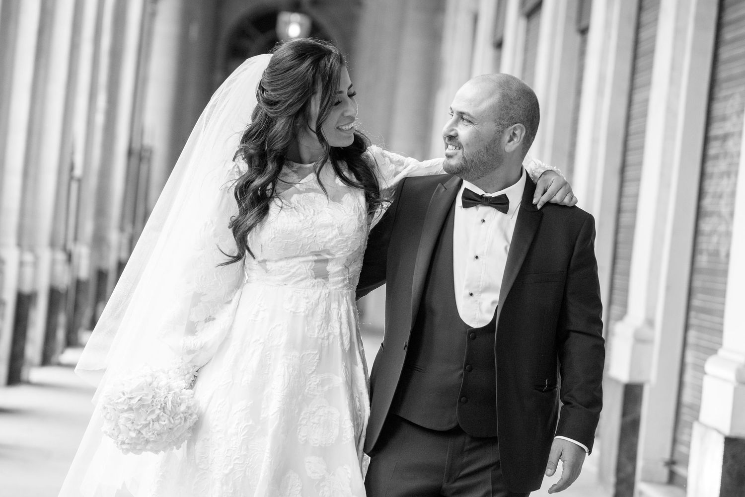 A bride and groom share a romantic moment while walking along a brick corridor with elegant arches.