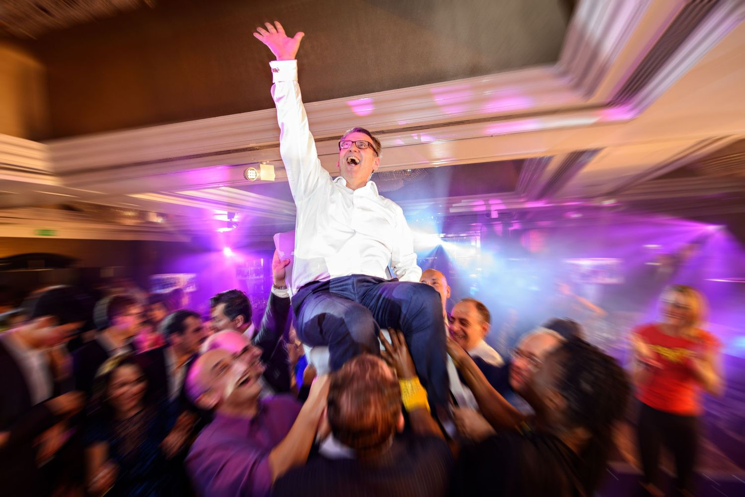 A crowded dance floor at a wedding reception with purple lighting and people dancing with hands raised in celebration.