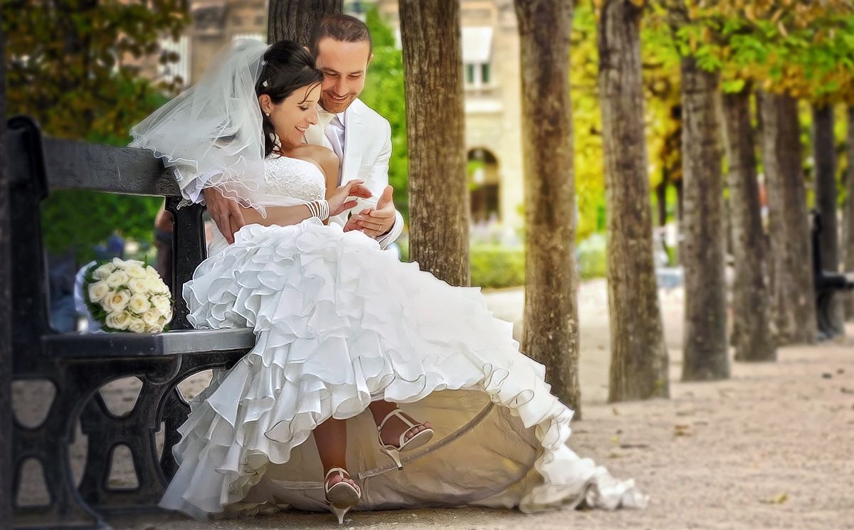Bride and groom sitting on a park bench, sharing an intimate moment in wedding attire among tree-lined path.