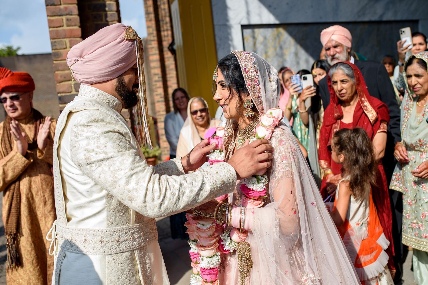 Sikh wedding ceremony with groom in pink turban exchanging garlands with bride in pink lehenga outdoors.
