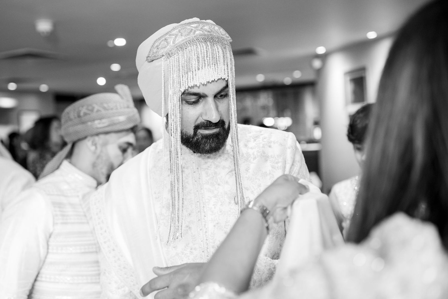 A groom in traditional Middle Eastern wedding attire with ornate beaded headpiece during a black and white ceremony moment.