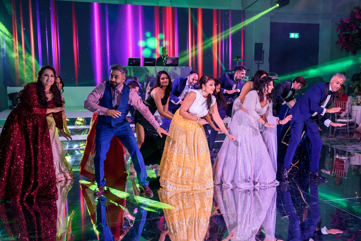 People in formal attire dance at a party with colorful neon lights and laser beams illuminating the dance floor.