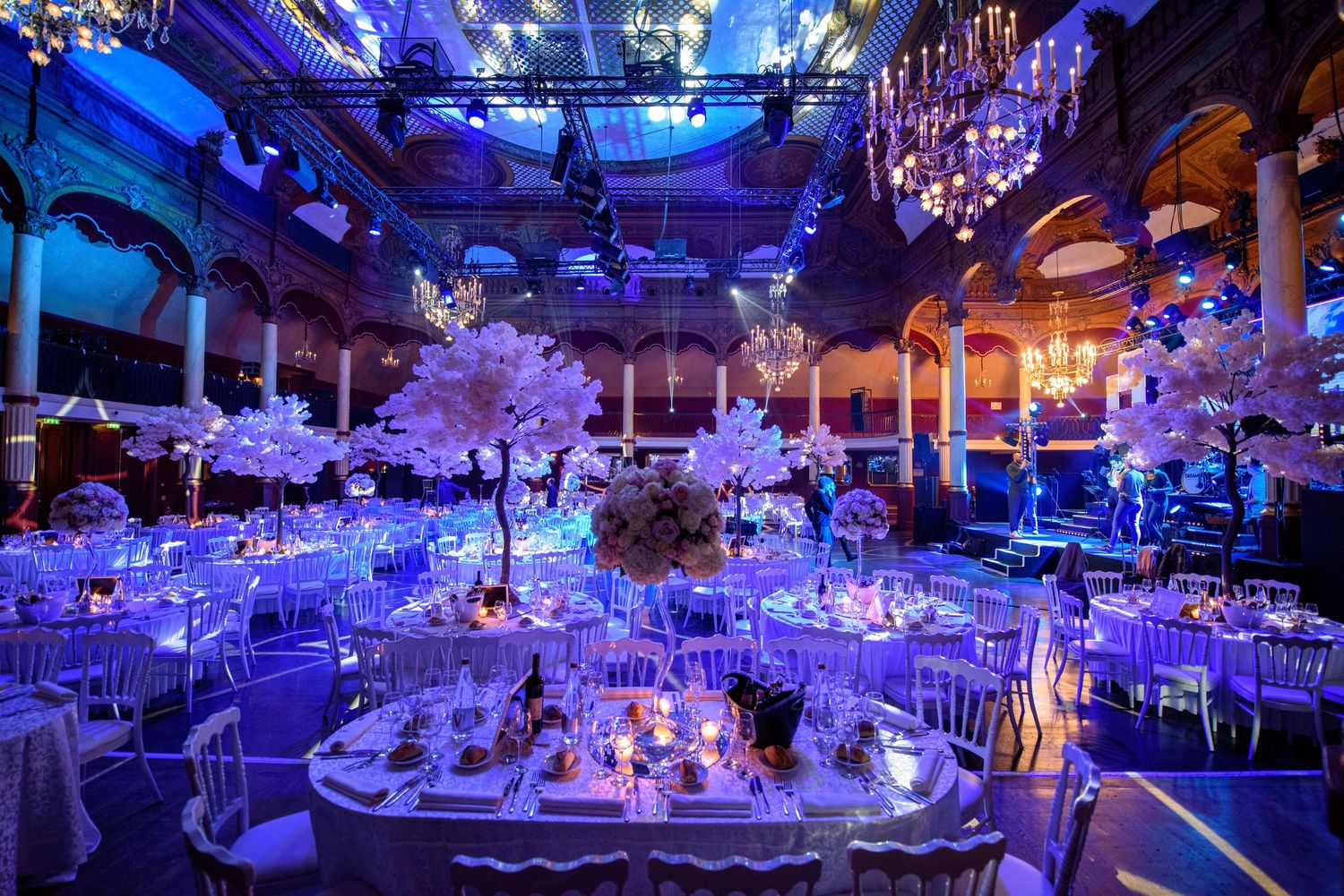 A luxurious ballroom decorated with blue uplighting, crystal chandeliers, and elegant table settings for a formal event.