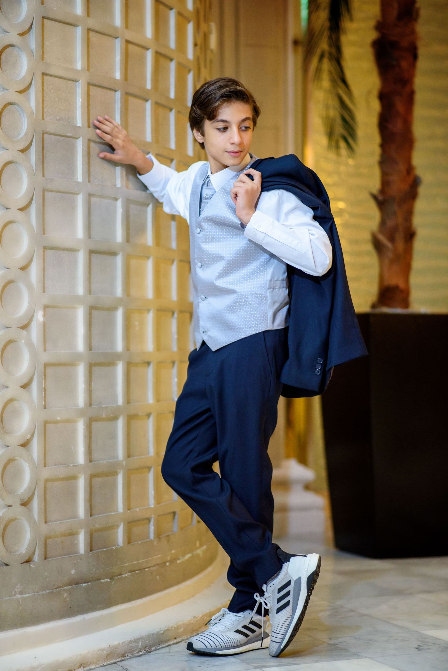 Young person in formal attire with vest and navy pants poses casually against decorative wall while holding jacket.