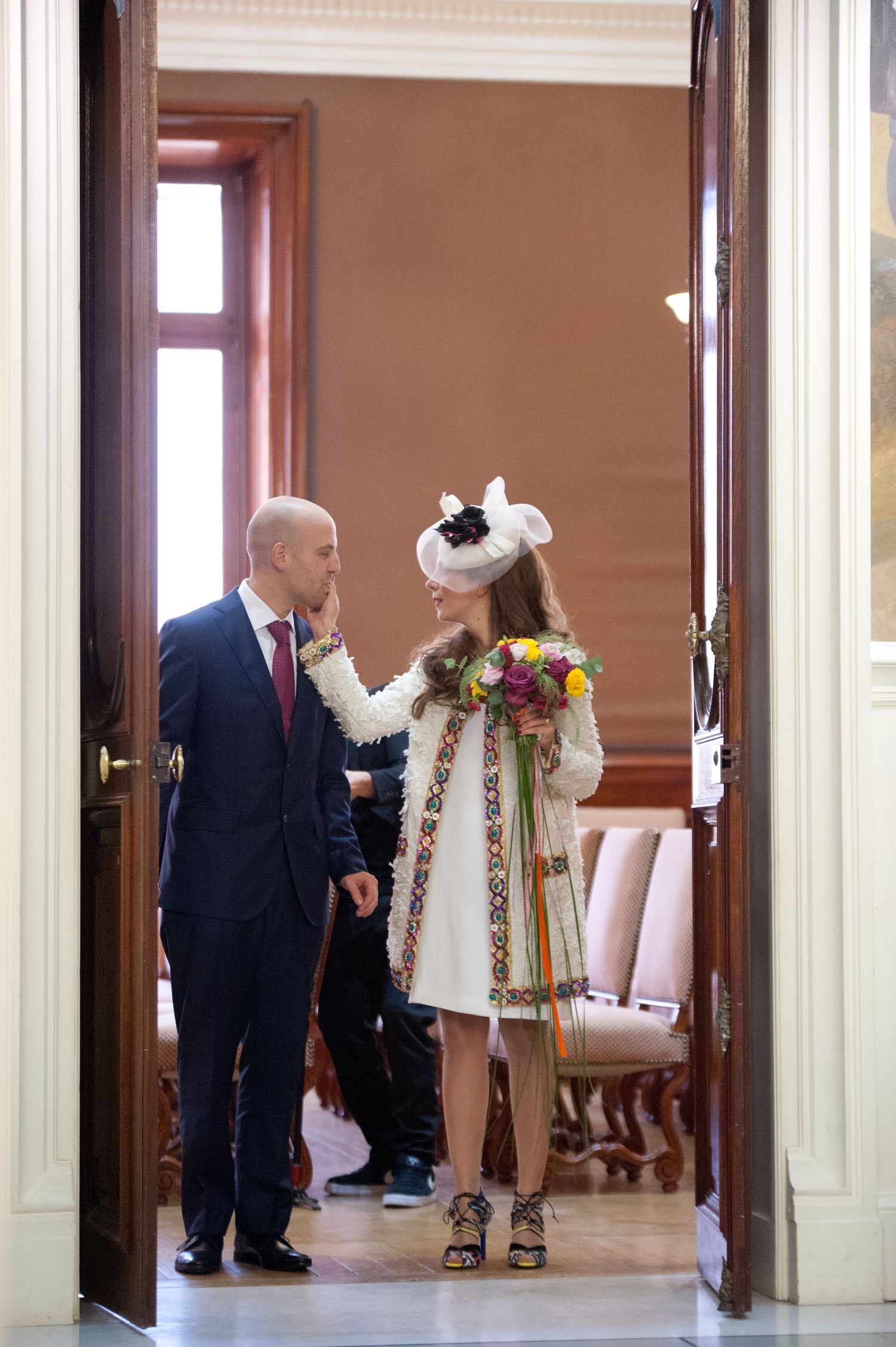 People in formal attire gather in an elegant hallway with wooden doors and cream-colored walls during a social event.