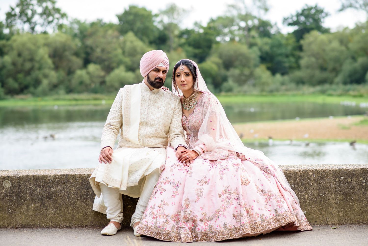 A Sikh couple in traditional wedding attire poses by a lake, the bride in pink lehenga and groom in cream sherwani.