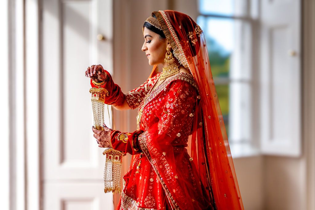 A bride in a red bridal lehenga and dupatta holds jewelry near a window, adorned in traditional gold ornaments.