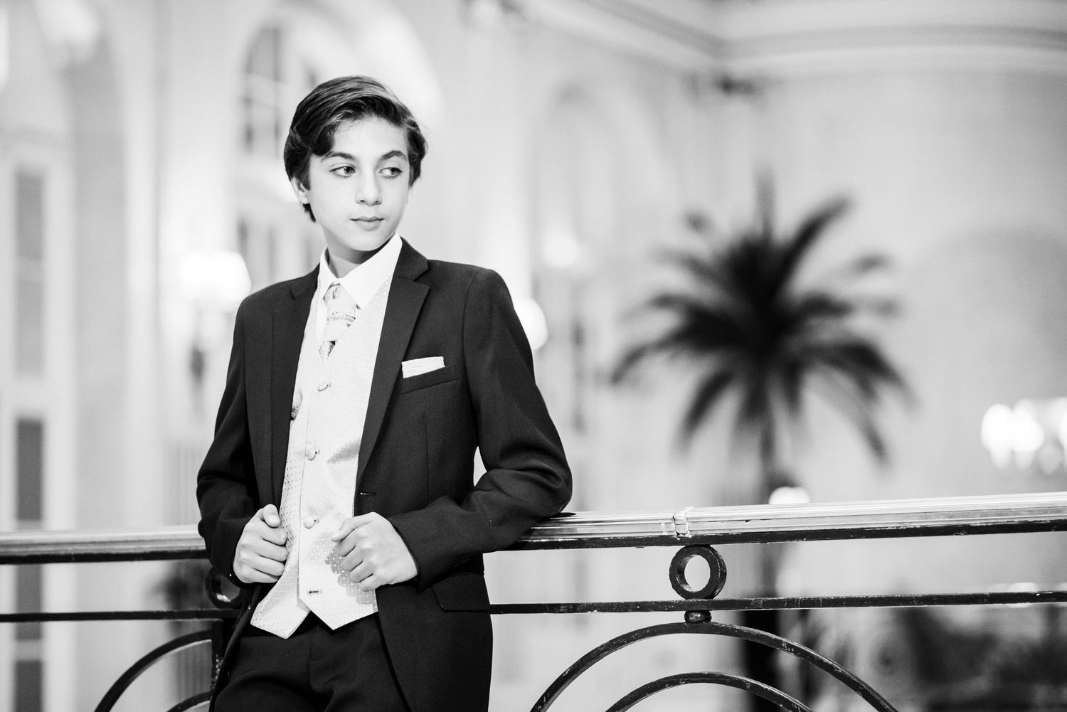 A black and white photo of someone in formal attire leaning against a railing with palm trees in the background.