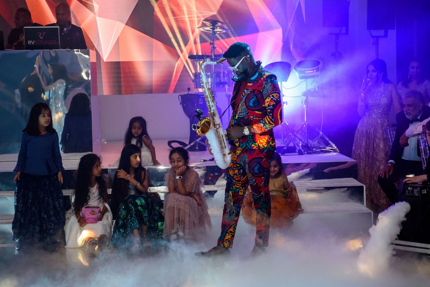 A performer in a colorful LED light suit entertains guests on a misty stage with dramatic purple and red lighting.