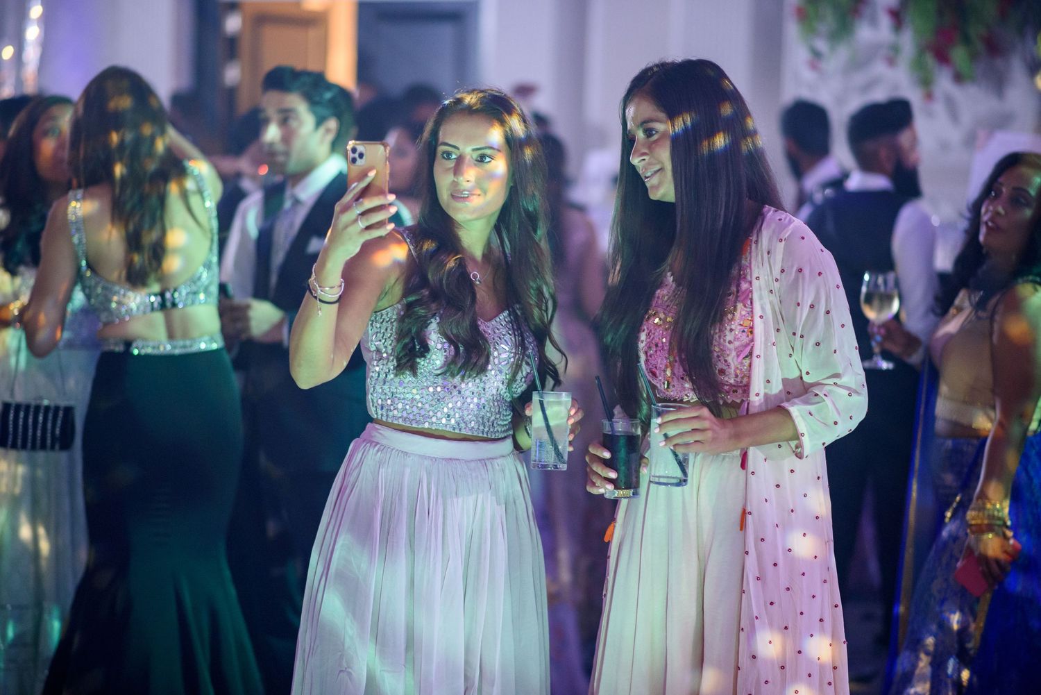 People in elegant dresses dancing and celebrating at a festive party with colorful lighting and decorations.